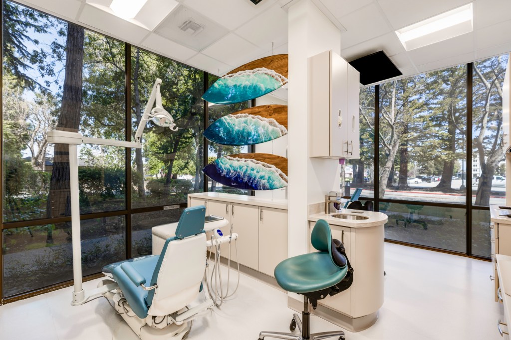 Bright dental operatory with teal-accented chair, ocean wave wall art, large windows showing trees outside, and modern equipment.