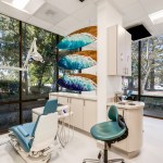 Bright dental operatory with teal-accented chair, ocean wave wall art, large windows showing trees outside, and modern equipment.