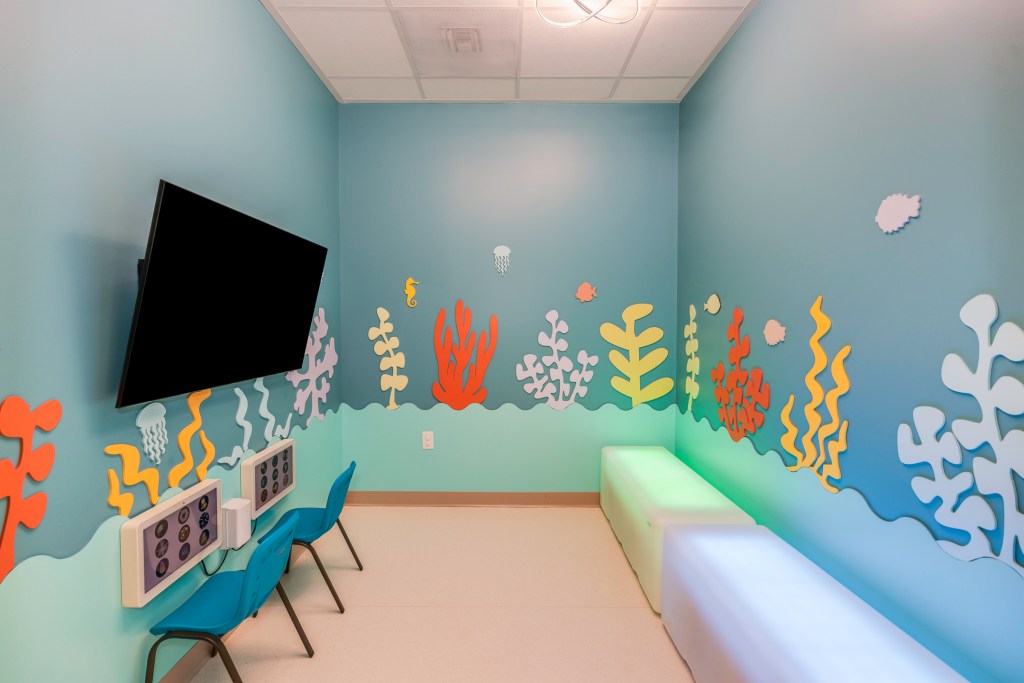 Child’s room with light blue walls decorated with colorful coral and sea life decals, a mounted TV, blue chairs, and a cushioned bench.