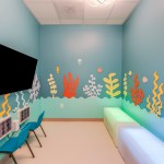 Child’s room with light blue walls decorated with colorful coral and sea life decals, a mounted TV, blue chairs, and a cushioned bench.