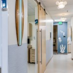 Clinic hallway with surfboard wall decor, sliding wood doors, and ocean-themed artwork under modern cloud pendant lights.
