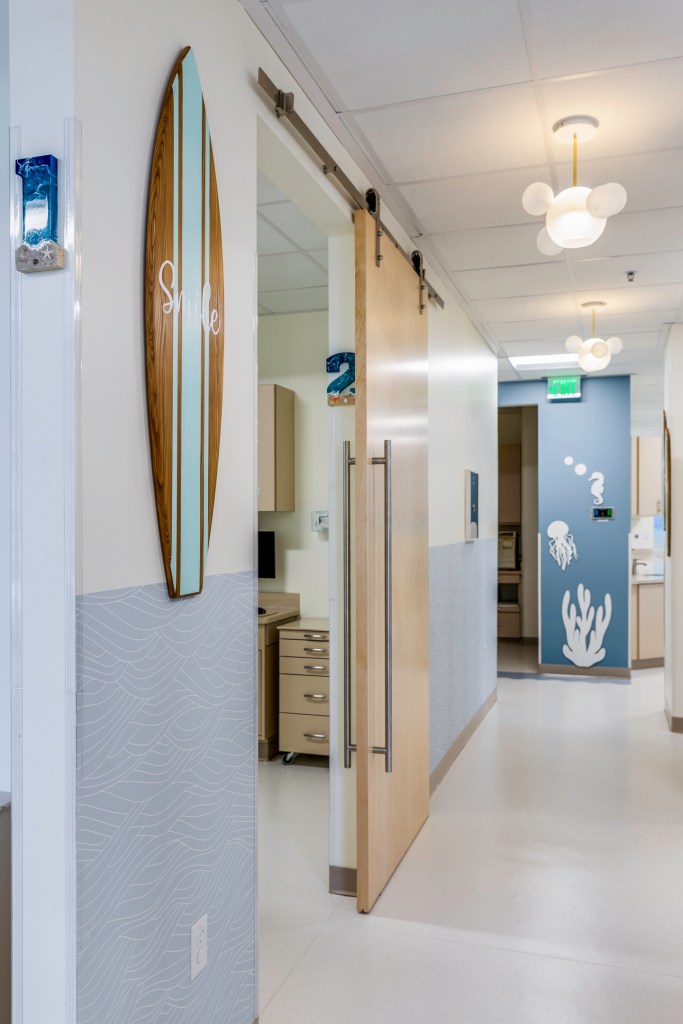 Clinic hallway with surfboard wall decor, sliding wood doors, and ocean-themed artwork under modern cloud pendant lights.