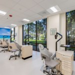 Dental treatment room with multiple operatories, ergonomic chairs, large windows overlooking greenery, and a beach mural on the back wall.