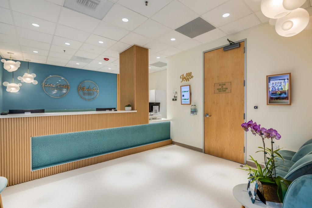 Wide view of clinic reception area showing the front desk, wooden door, digital screen, and potted orchids near seating.
