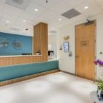 Wide view of clinic reception area showing the front desk, wooden door, digital screen, and potted orchids near seating.