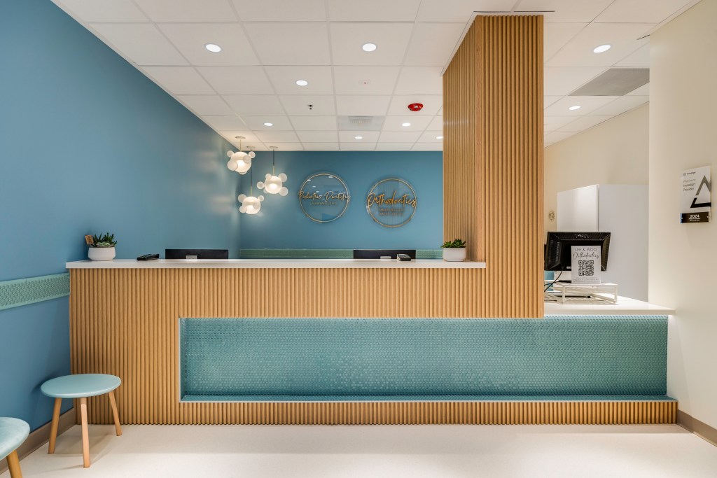 Modern dental reception desk with light wood slats and teal accents, under bright ceiling lights and a blue accent wall.