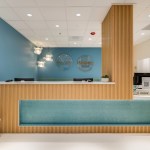 Modern dental reception desk with light wood slats and teal accents, under bright ceiling lights and a blue accent wall.