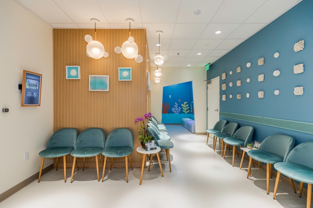 Waiting area with rows of teal chairs, wood-paneled walls, hanging cloud lights, and colorful underwater-themed hallway in background.
