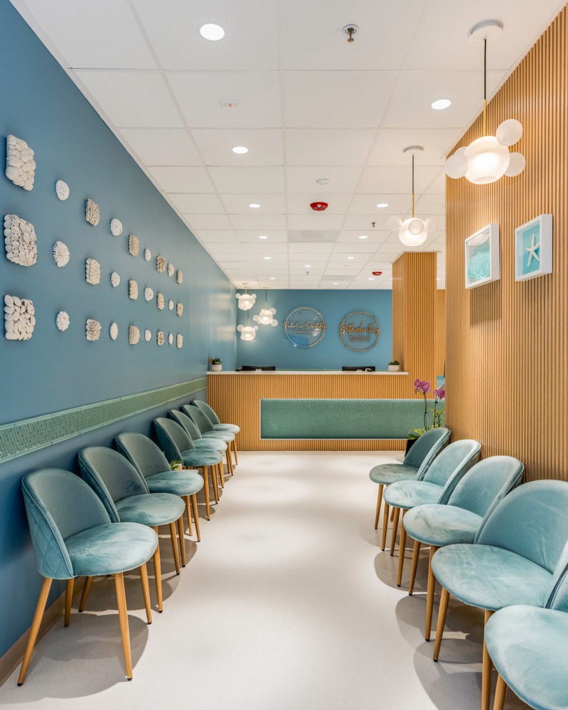 Symmetrical view of waiting room with teal chairs lined along both sides, leading to reception desk under soft pendant lighting.