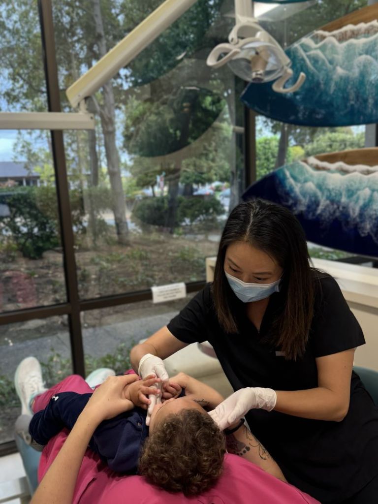 Dental professional wearing mask and gloves gently examining a child’s teeth in a bright operatory with ocean-themed wall art visible through the window.