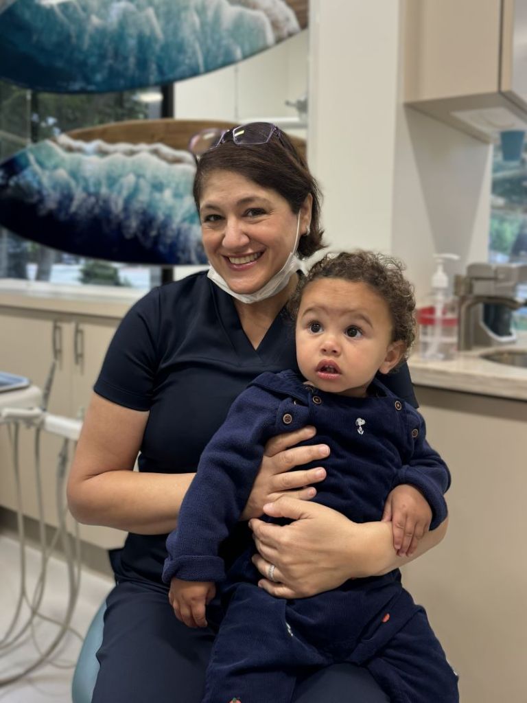 Smiling dental professional holding a young child in a clinic setting, with ocean-themed wall art in the background.