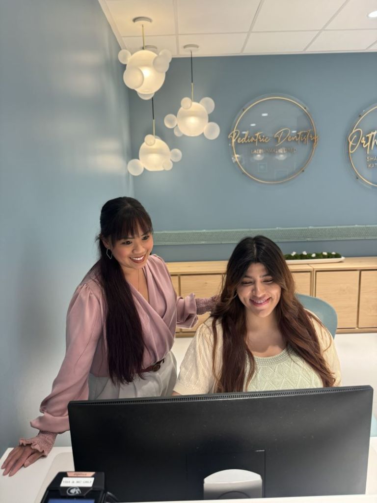 Two team members at a reception desk — one leaning over to assist another seated at a computer, both smiling.