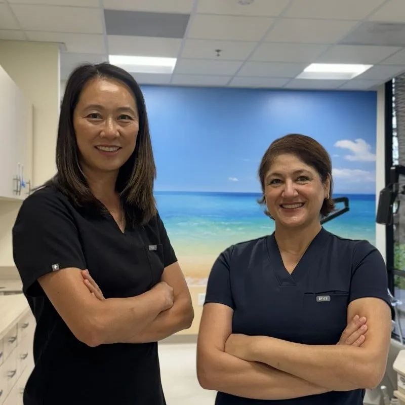 Two smiling female dental professionals in scrubs standing with arms crossed in a clinic, with an ocean mural behind them.