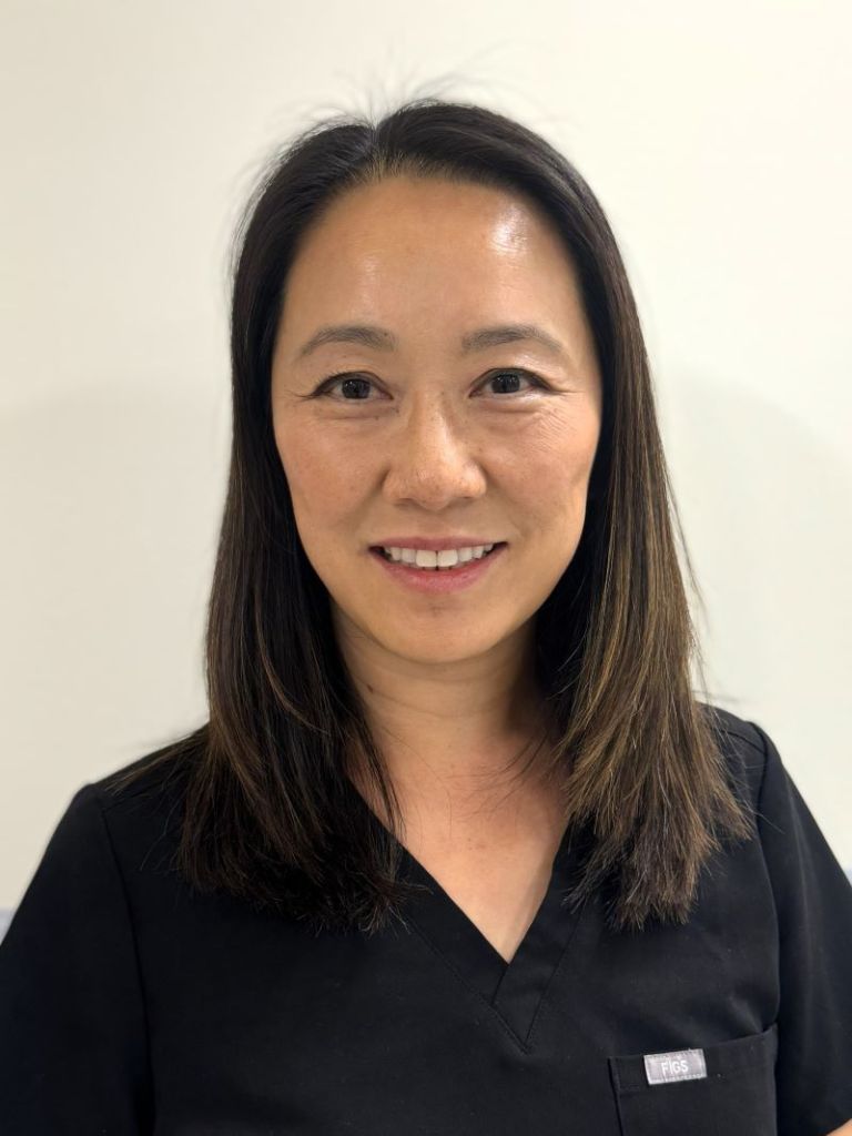 Headshot of a smiling female dental professional with shoulder-length dark hair, wearing black scrubs against a plain white background.