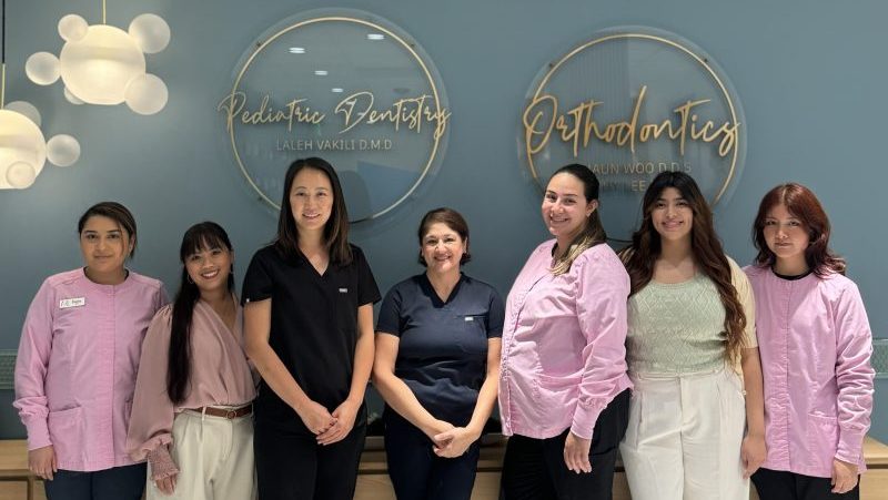 Dental team of six smiling staff members standing in front of a blue wall with “Pediatric Dentistry” and “Orthodontics” signage.