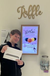 Child smiling while holding a gift box in front of a “Raffle” sign and digital display promoting a “Winter Raffle” with colorful prizes.